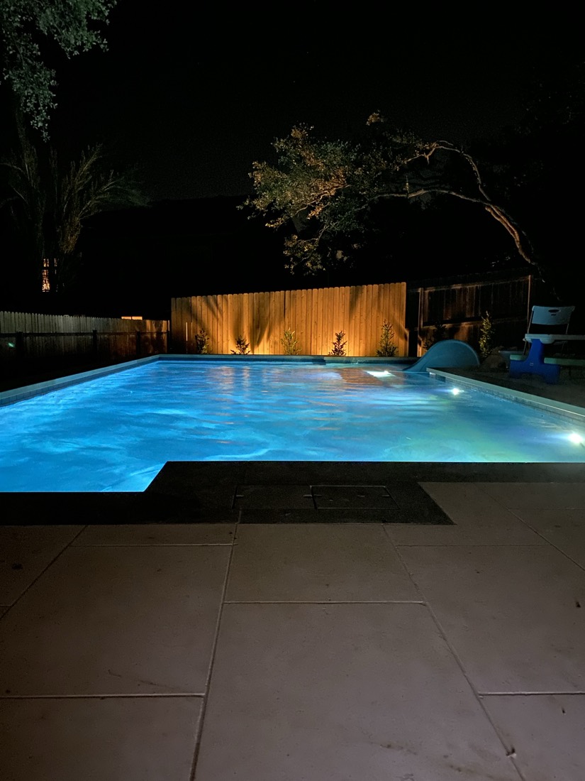 A pool glowing blue at night, slide at the far end, cedar fence lit warm behind it.